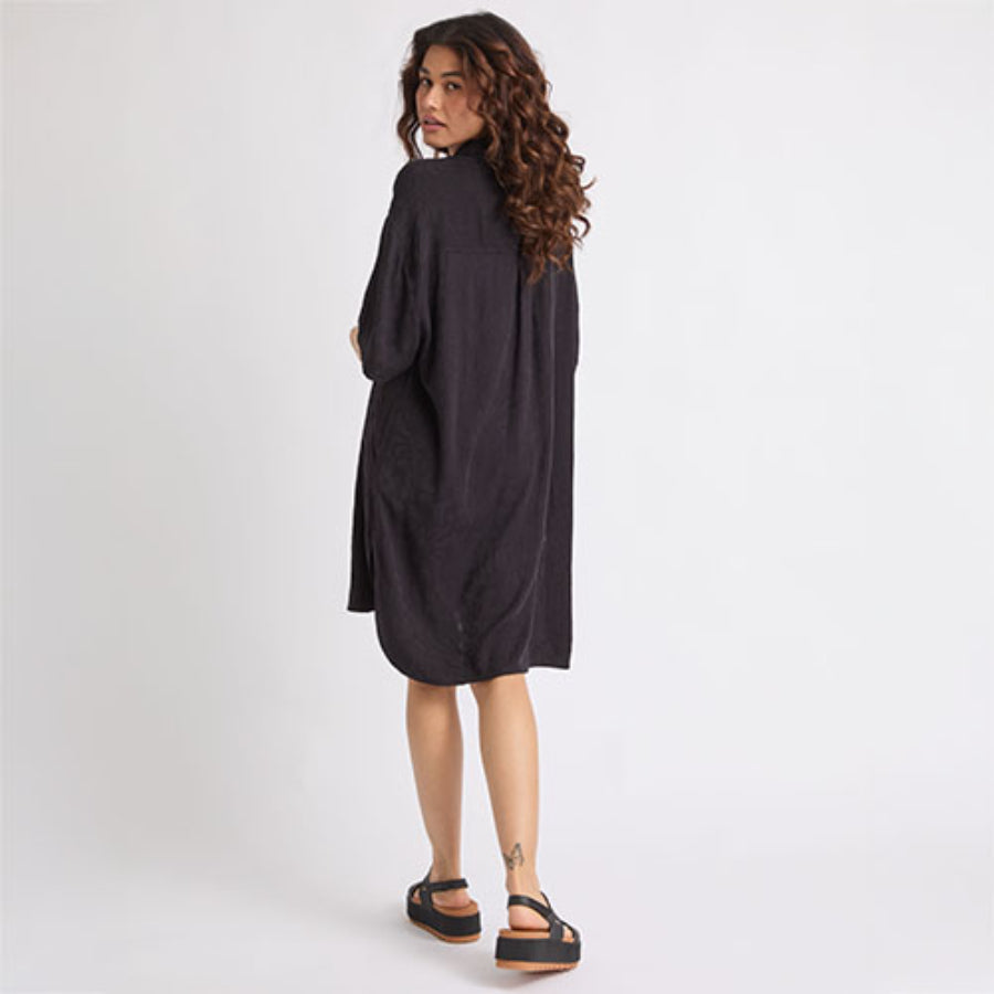 Woman wearing a black Ollie Shirt Dress seen from back, paired with black platform sandals facing right, surf-inspired comfort style.
