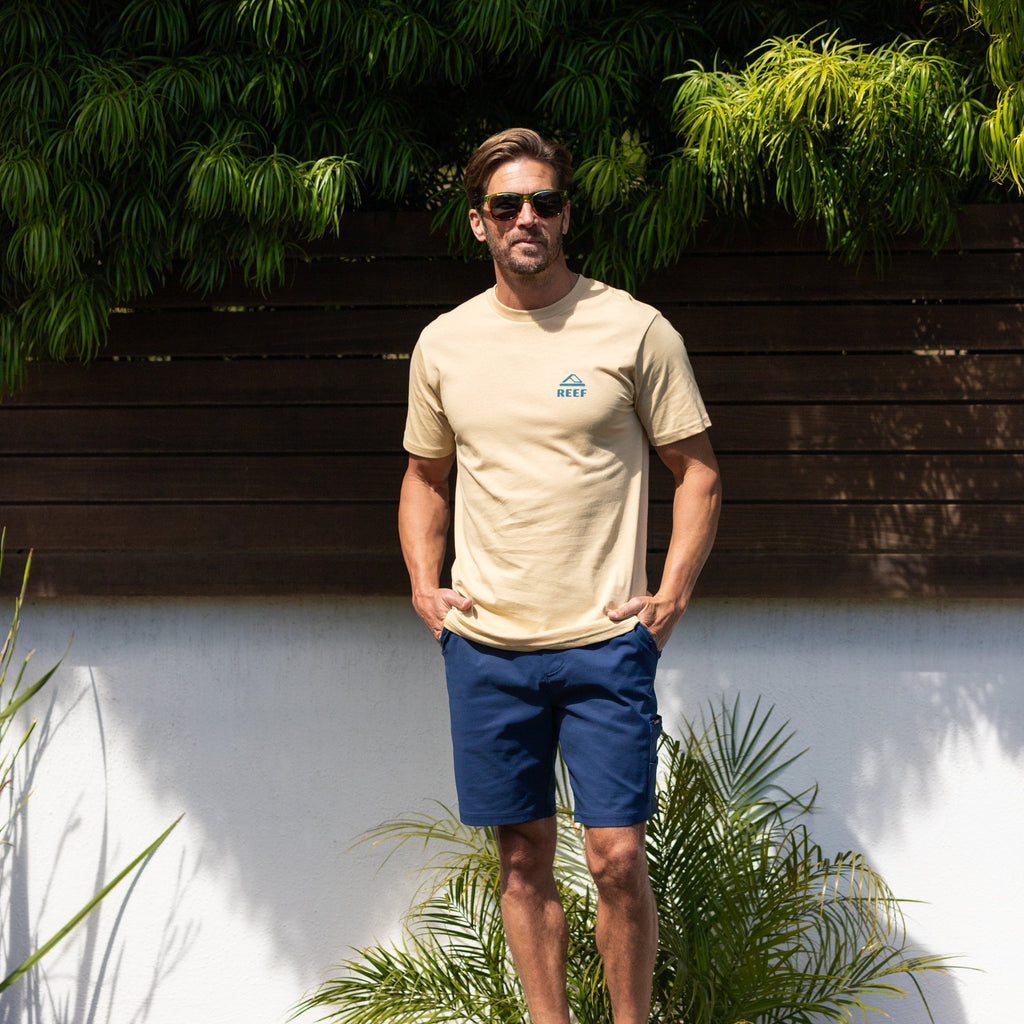 Man wearing a beige Xavier Tee with sunglasses, standing outdoors by plants and wooden fence in bright sunlight