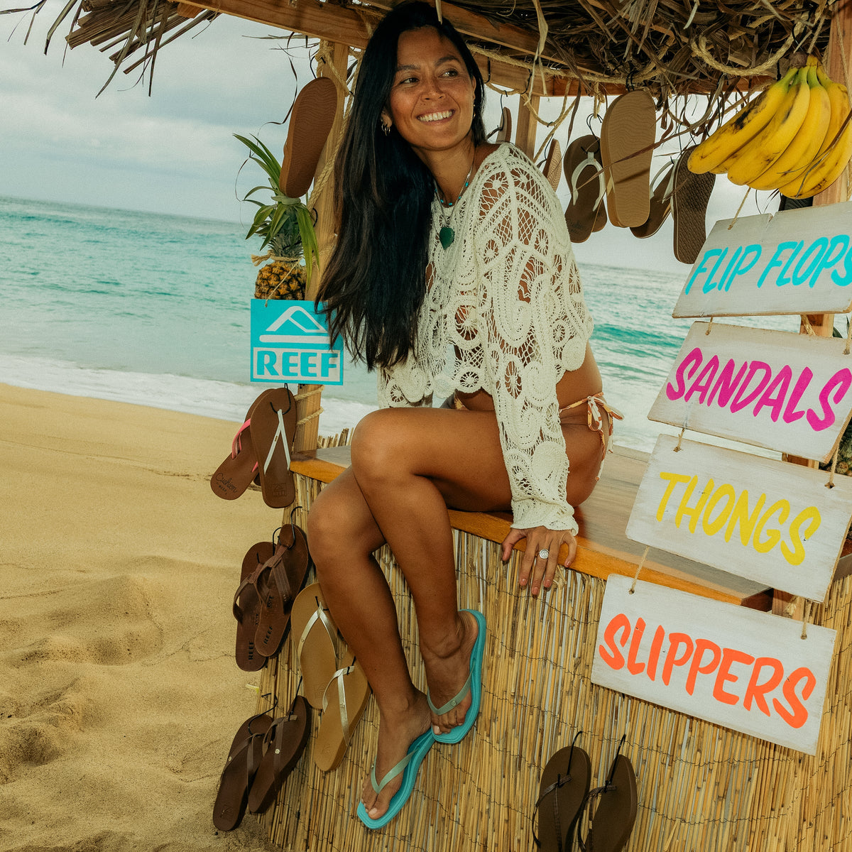 Woman wearing Solaye teal sandals facing slightly left, sitting on beach hut counter with surf-inspired footwear displayed nearby.