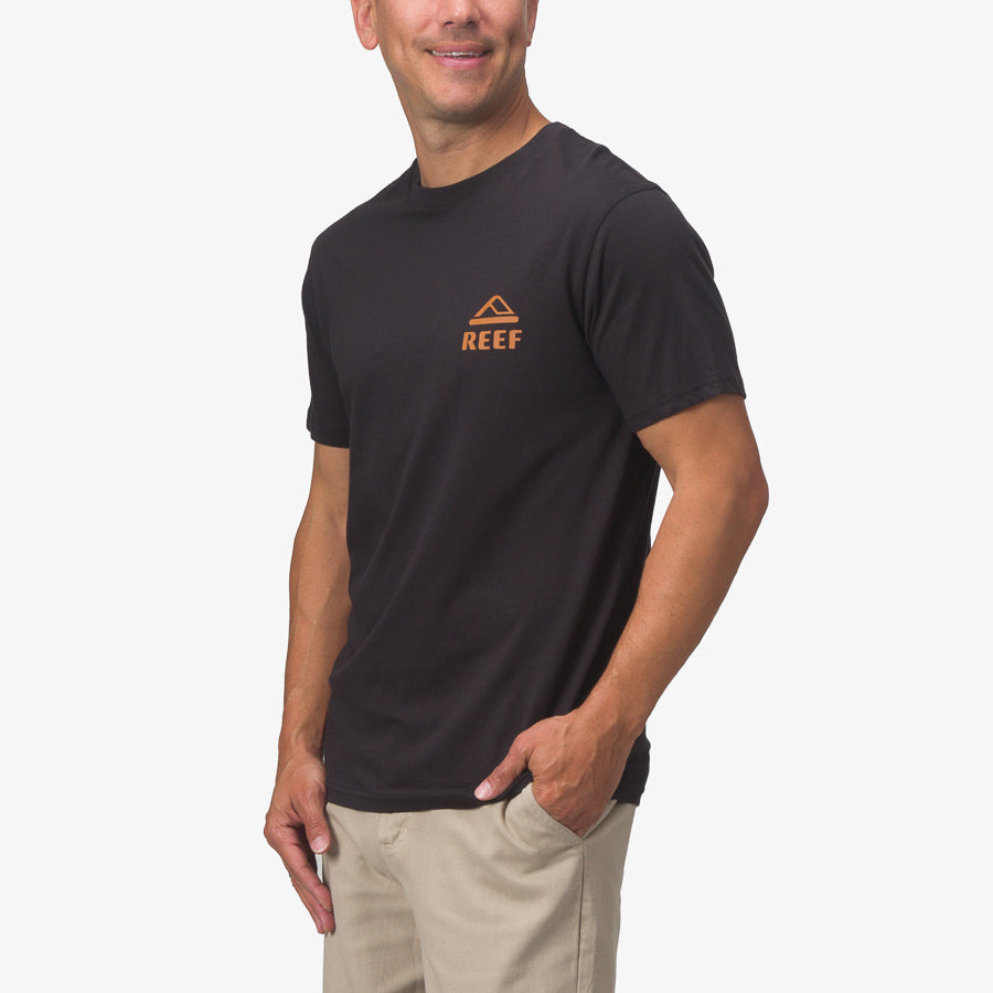 Man wearing a black Wayne Tee with orange REEF logo, shown from front right side, comfortable beach-inspired apparel.